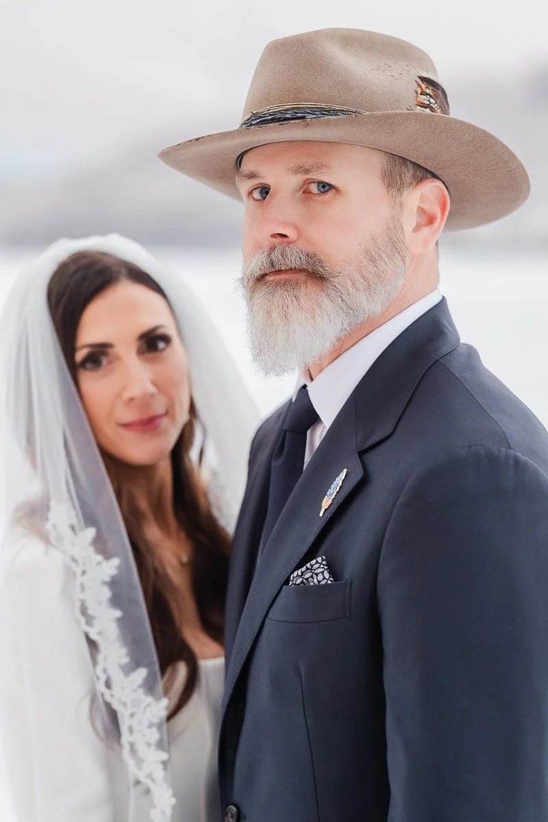 A bride in a lace veil and a groom in a suit with a feathered hat pose outdoors. The snowy background creates a serene, romantic atmosphere.