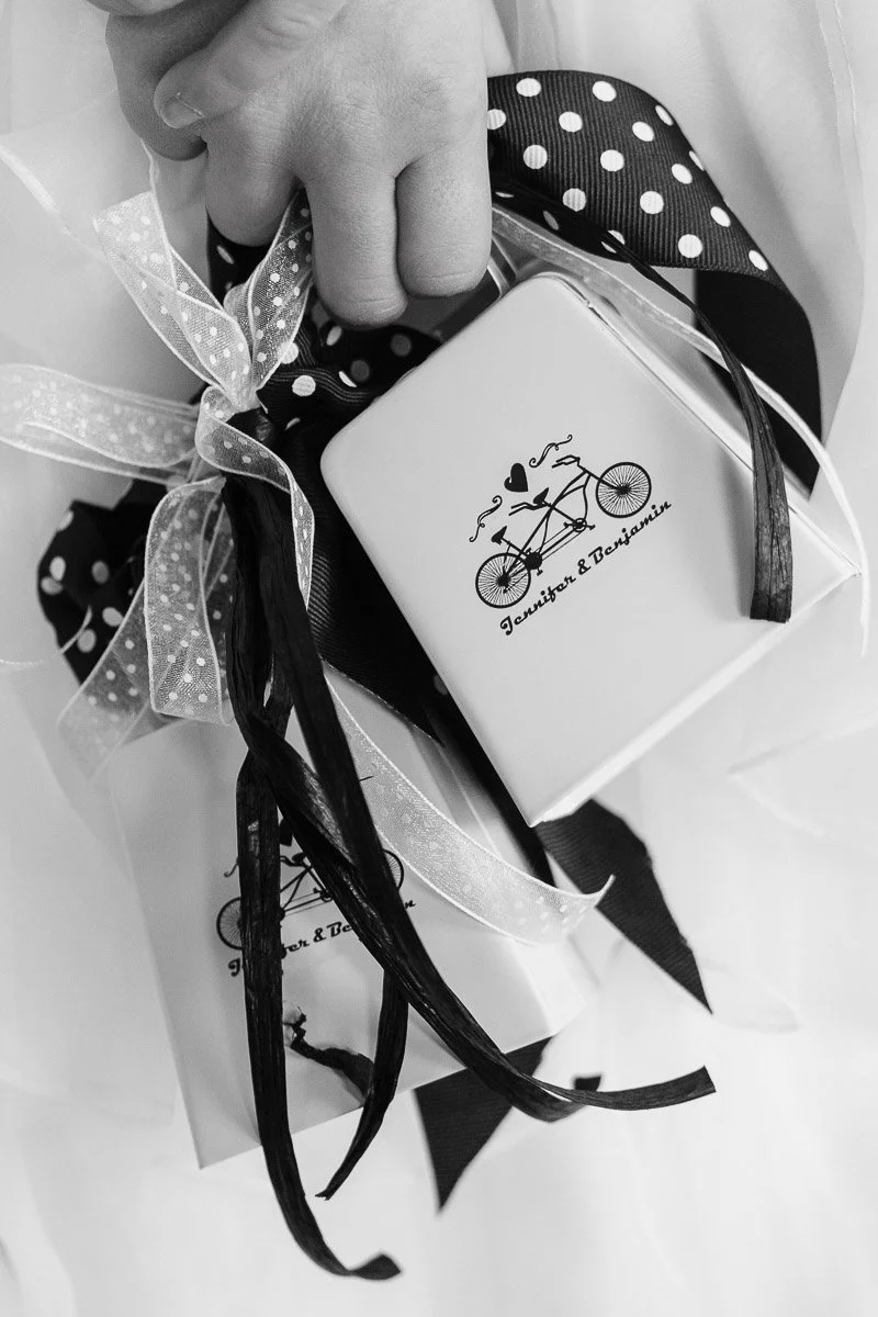 A child's hand holding a gift box adorned with black and white polka dot and sheer ribbons. Illustration of a tandem bike with hearts adds charm.