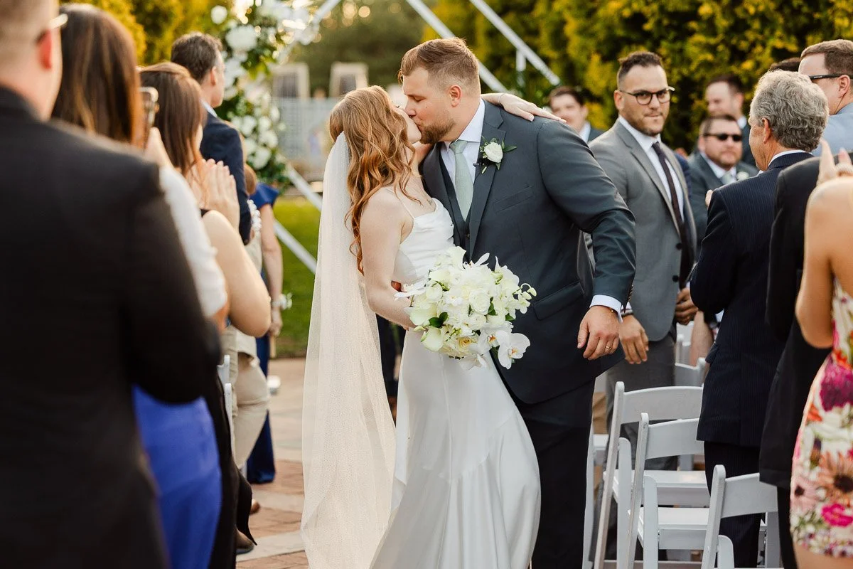Bride and groom sharing a kiss at their wedding ceremony, surrounded by applauding guests. The bride holds a bouquet, exuding joy and romance.