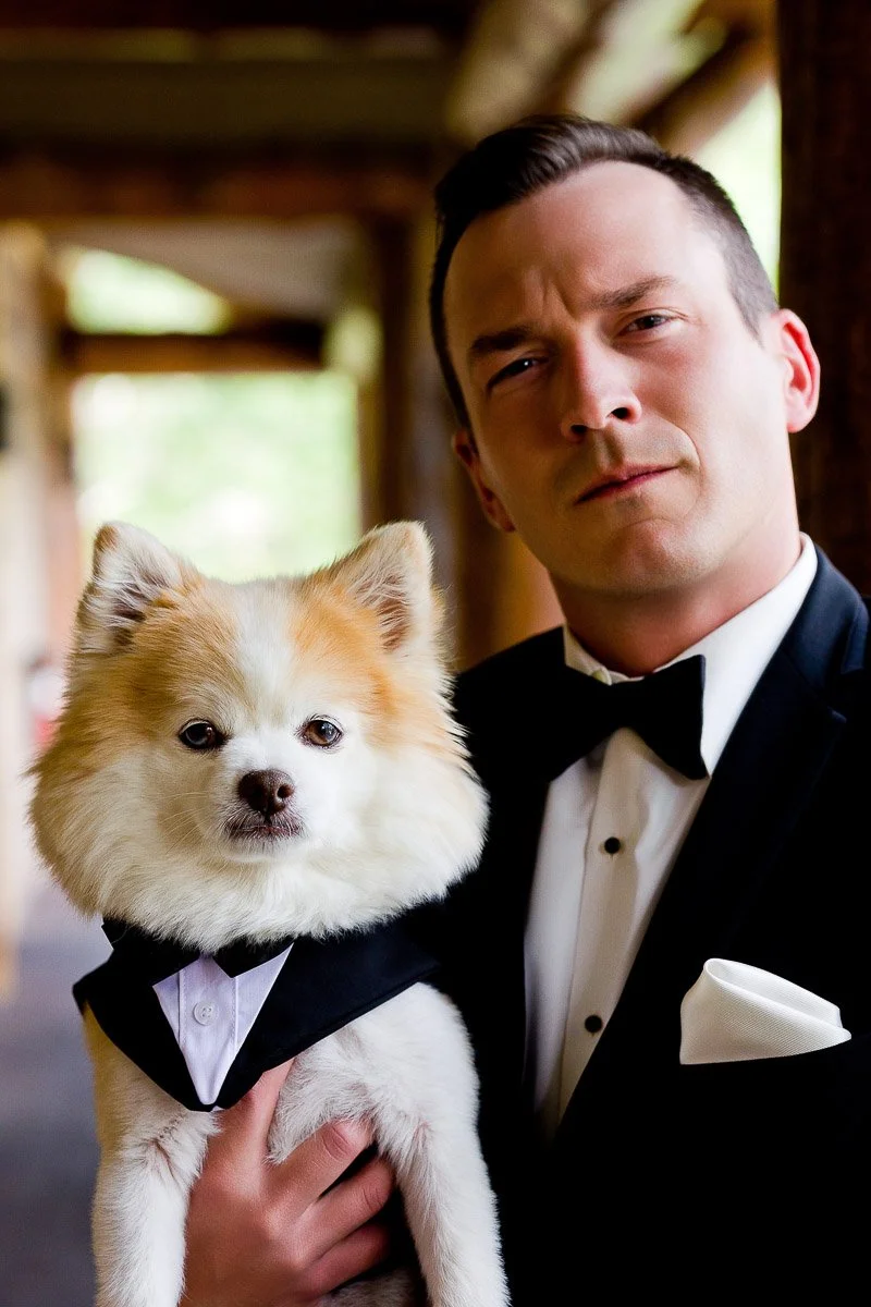 A man in a tuxedo confidently holds a small fluffy dog also dressed in a tuxedo. They stand in an elegant setting, both looking intently at the camera.