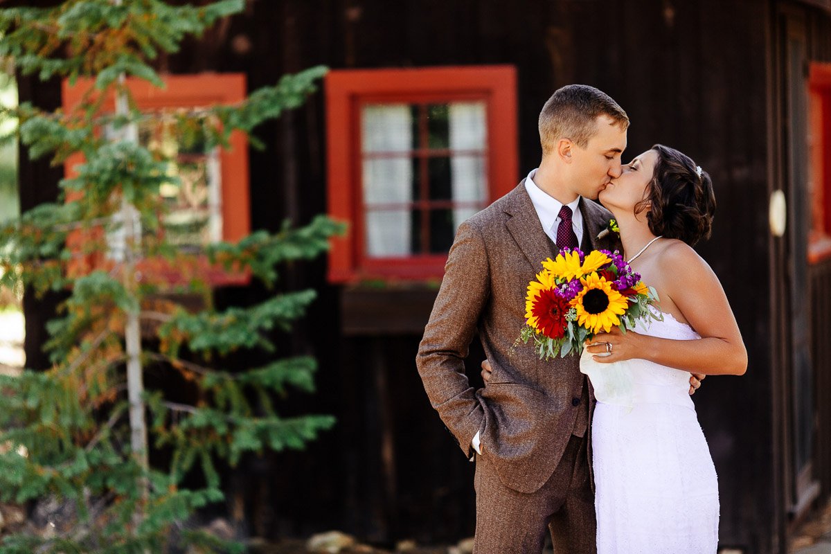 A couple kisses in front of a rustic cabin. The bride holds a vibrant bouquet with sunflowers. The mood is joyful and romantic captured by YMCA of the rockies wedding photographer tomKphoto