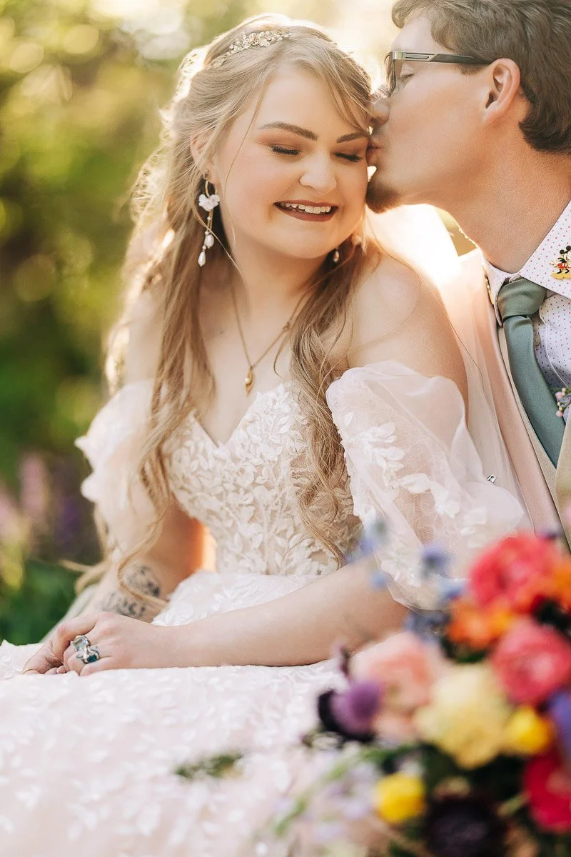 Bride in lace gown smiles as groom kisses her forehead, surrounded by vibrant flowers and lush greenery. The scene radiates joy and romance.
