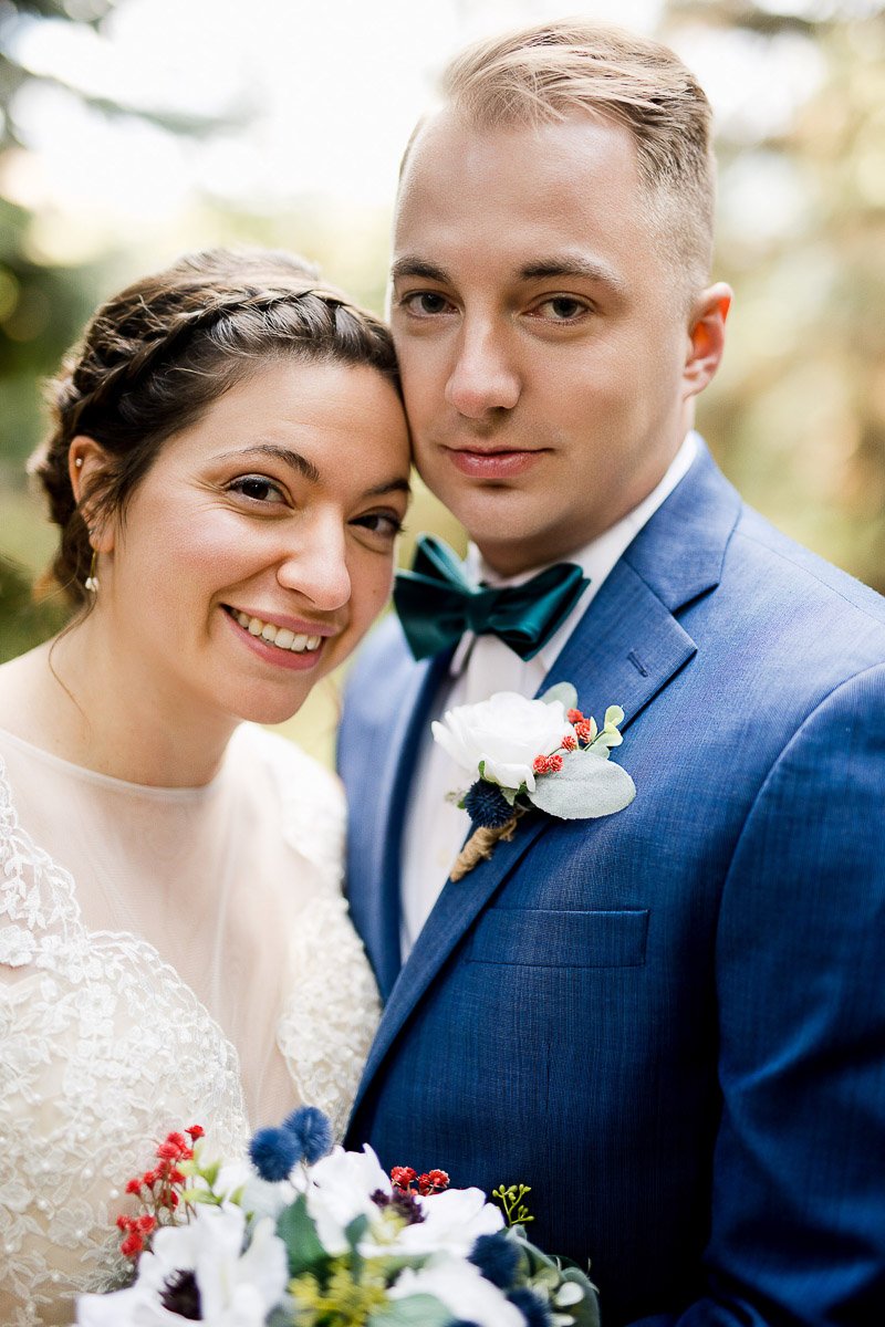 A bride in a lace dress smiles while leaning her head against a groom in a blue suit. He wears a teal bow tie and boutonniere. Both look joyful.