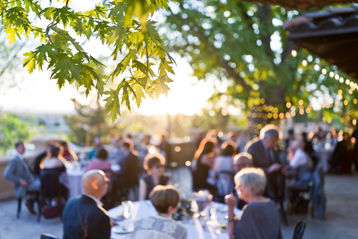 Outdoor gathering at sunset with people seated at tables under string lights. Green leaves in the foreground and a warm, festive atmosphere during a Villa Parker wedding in Parker, Colorado