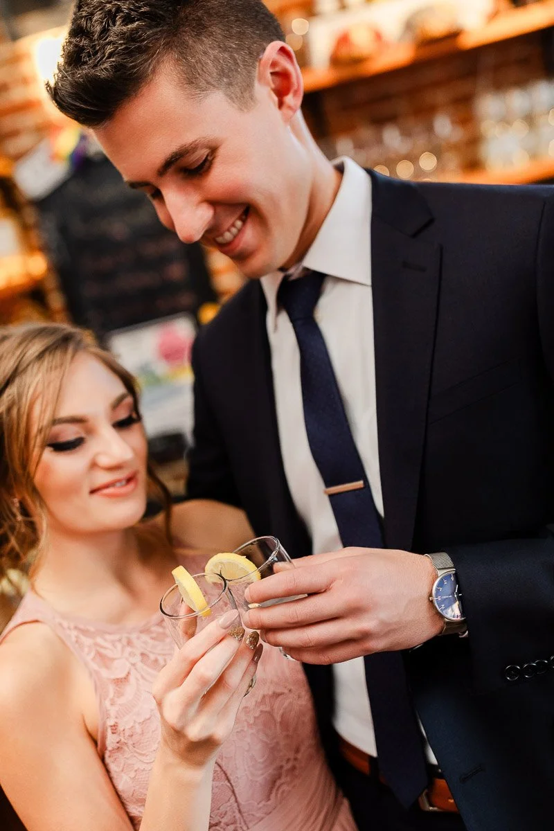 A smiling couple clinks glasses with lemon wedges. The woman wears a pink lace dress, and the man is in a dark suit. The setting is warm and inviting.