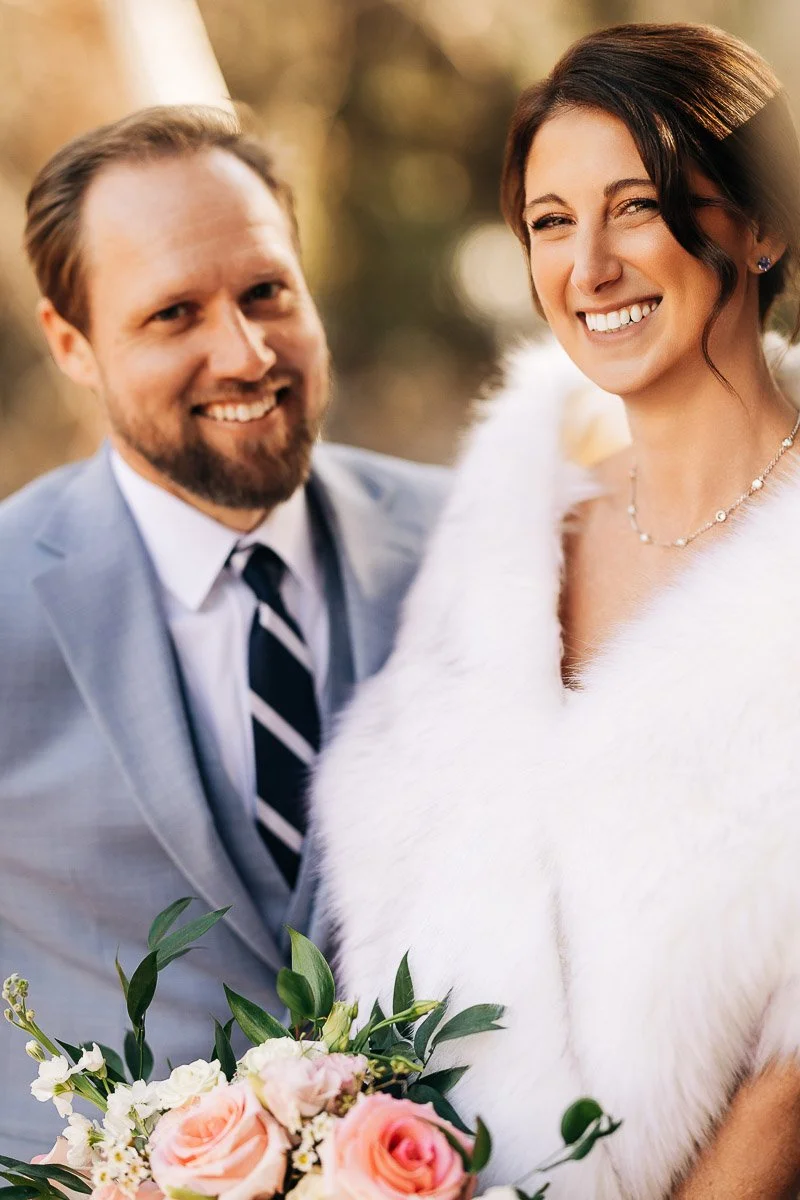 A smiling couple on their wedding day; the bride wears a white fur shawl and holds a bouquet of pink roses, while the groom is in a light gray suit.