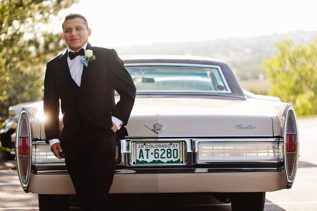 Young man in a tuxedo leans confidently against a vintage Cadillac with a Colorado license plate, with sunlight creating a warm glow. Background: blurred greenery and clear sky.