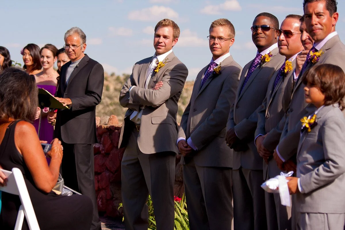 A group of groomsmen in gray suits with yellow boutonnieres stand facing a seated woman outdoors. Sunlight illuminates the scene, creating a joyful, formal atmosphere.