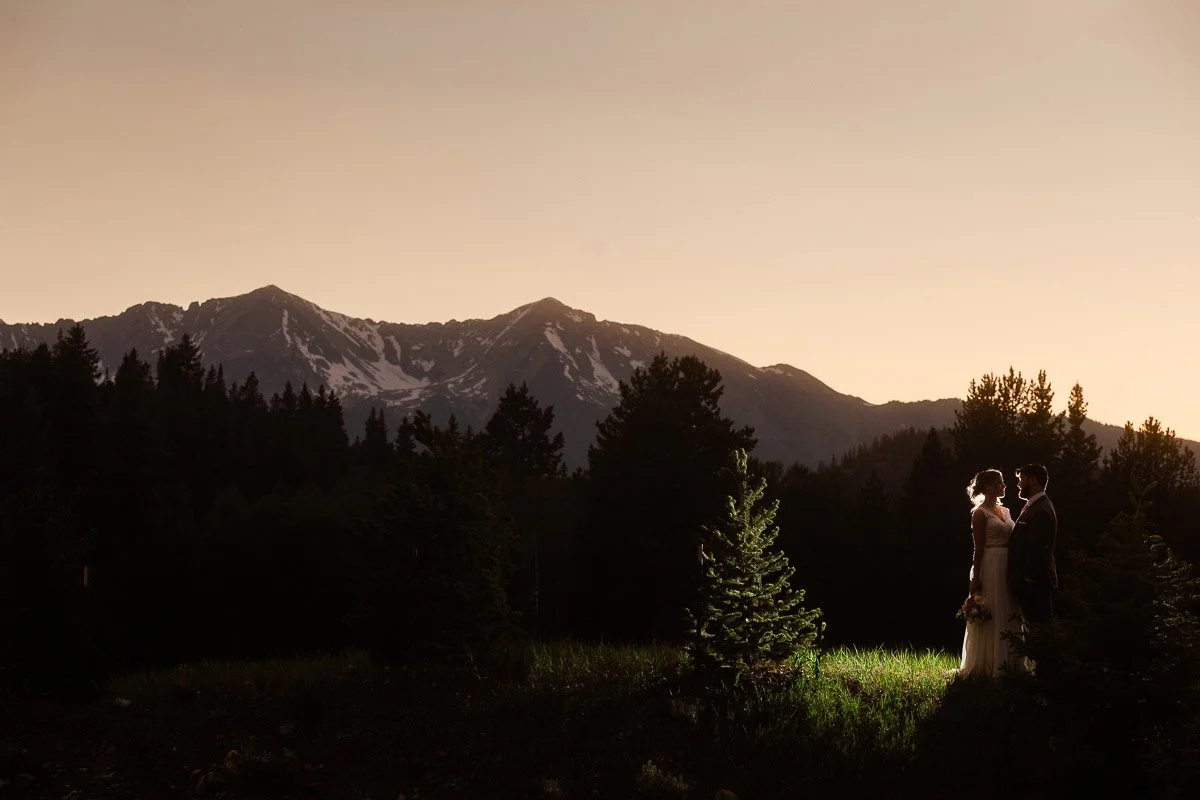 A bride and groom stand together in a sunlit clearing against a backdrop of towering mountains and pine trees at dusk, creating a serene, romantic scene.