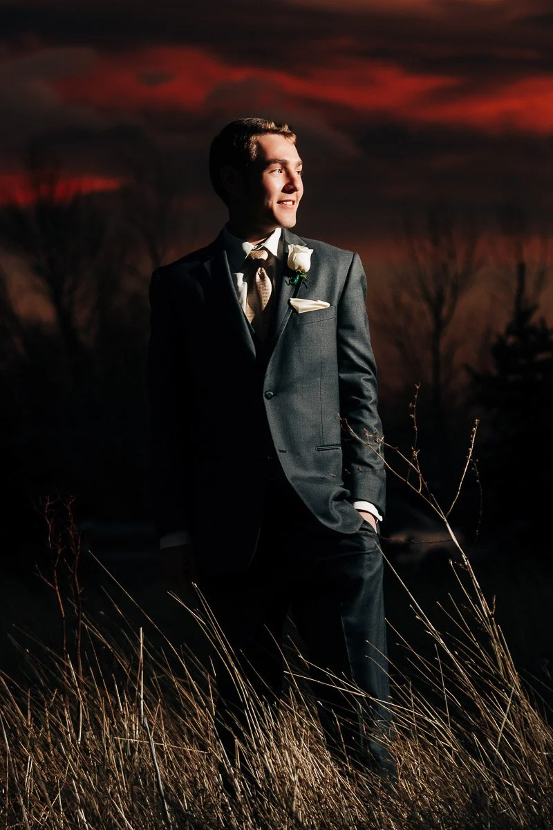 A man in a dark suit stands confidently in tall grass under a dramatic, red sunset sky, exuding elegance and hope as he gazes to the side.