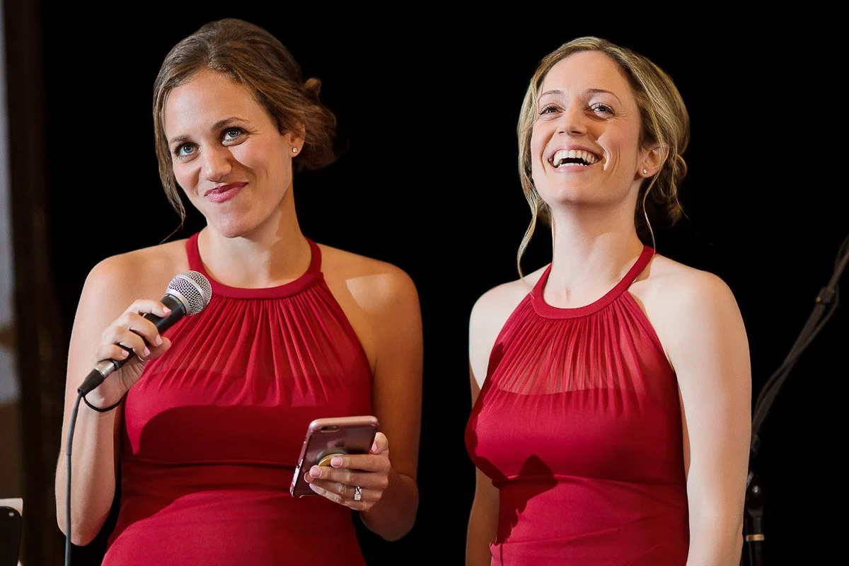 Two women in matching red dresses stand together. One holds a microphone and phone, smiling softly, while the other laughs, both exuding joy and camaraderie.