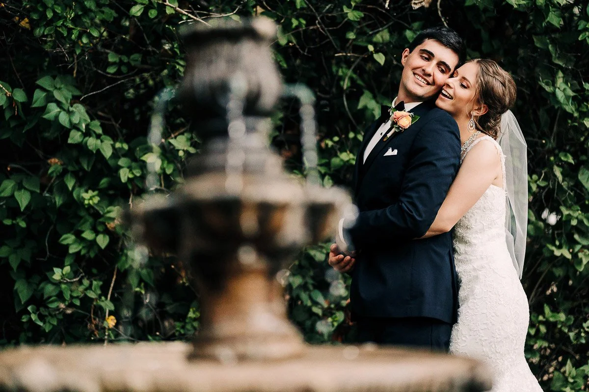 A joyful bride and groom embrace beside a fountain with lush greenery in the background, conveying romance and happiness in an outdoor wedding setting.