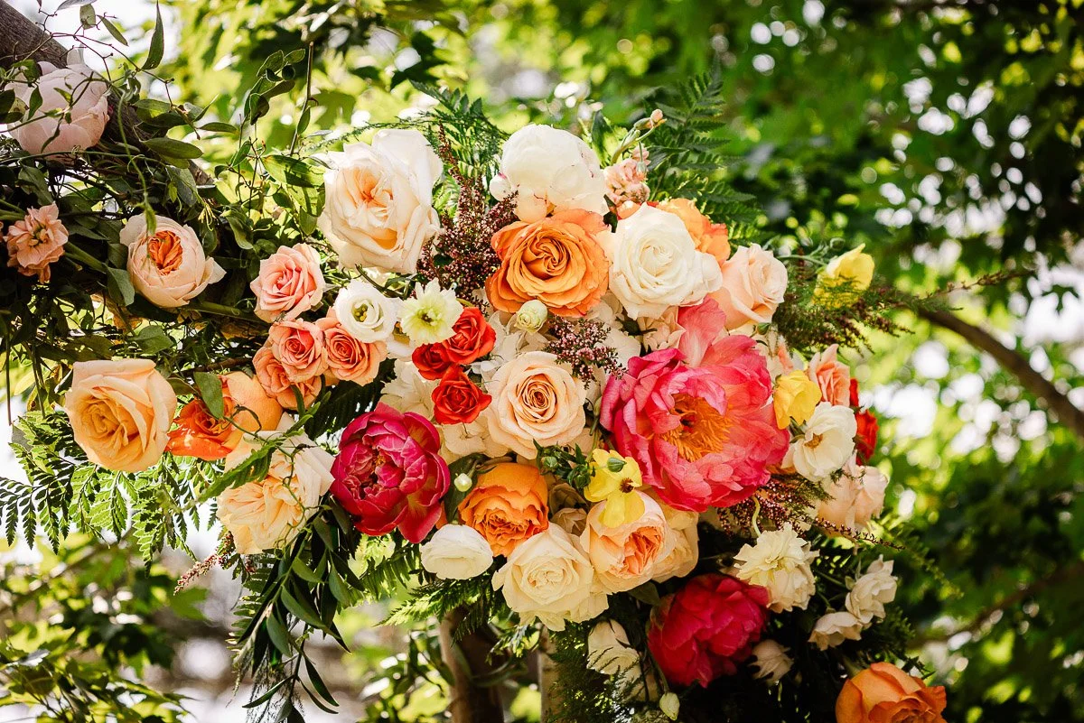 A vibrant floral arrangement with roses in shades of peach, pink, orange, and white, surrounded by green foliage. Sunlight filters through leafy trees.
