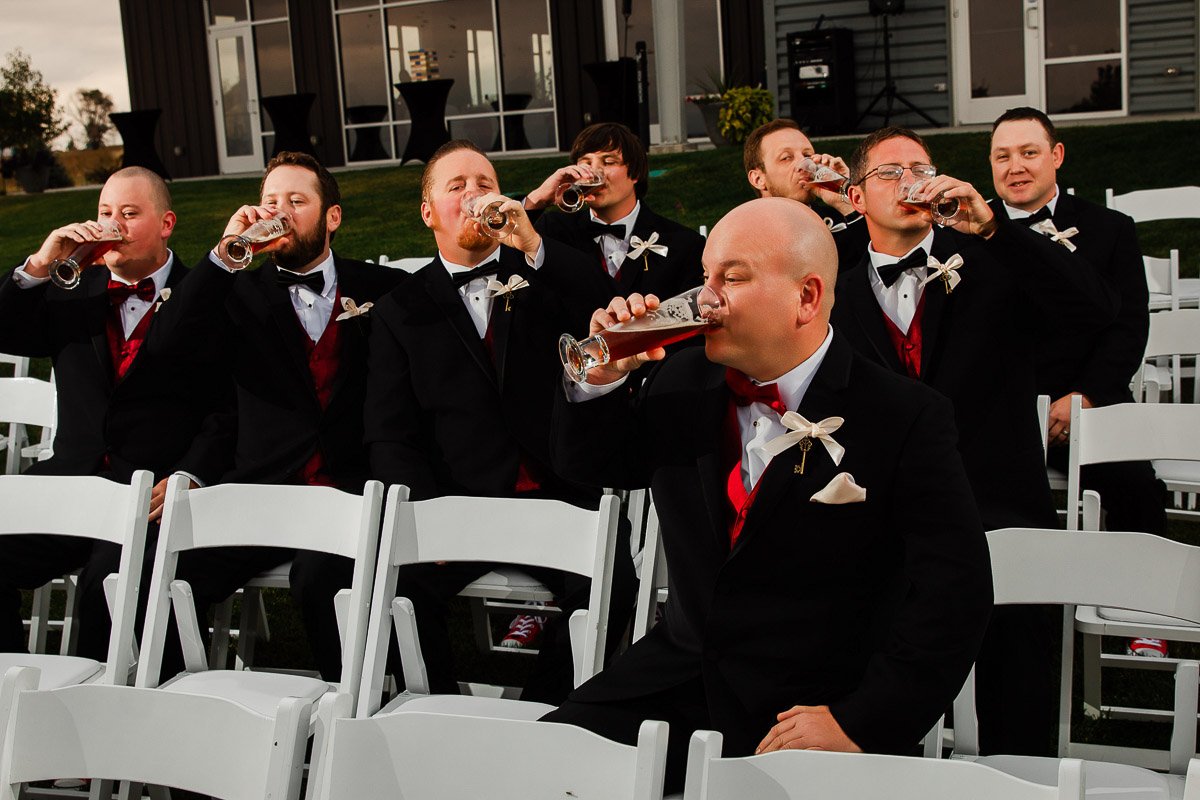 A group of groomsmen in black tuxedos with red vests drink beer from tall glasses while seated on white chairs outdoors, conveying a celebratory mood.