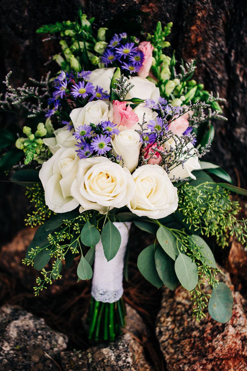 A vibrant bouquet with white roses, pink buds, and purple flowers, surrounded by green leaves, rests against a textured tree trunk and rocks.