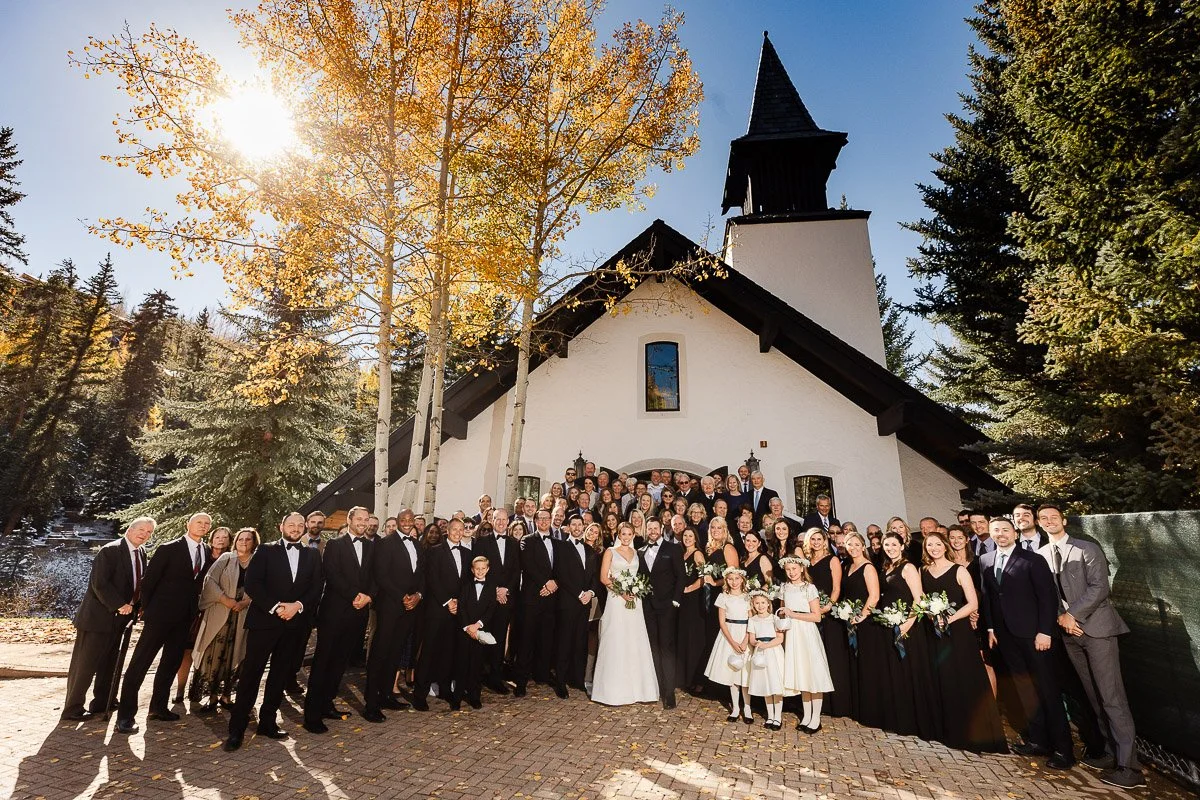 A large wedding group poses joyfully in front of a quaint chapel surrounded by autumn trees. The sun shines brightly, casting a warm, cheerful mood.