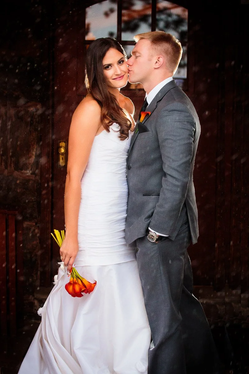 A bride in a white gown holds orange flowers, smiling softly. The groom, in a gray suit, kisses her cheek. Warm lighting and a dark wooden door backdrop set a romantic tone.