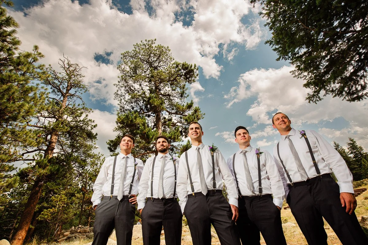 Five men in white shirts with suspenders stand smiling under a blue sky with scattered clouds, surrounded by tall pine trees. The mood is joyful.