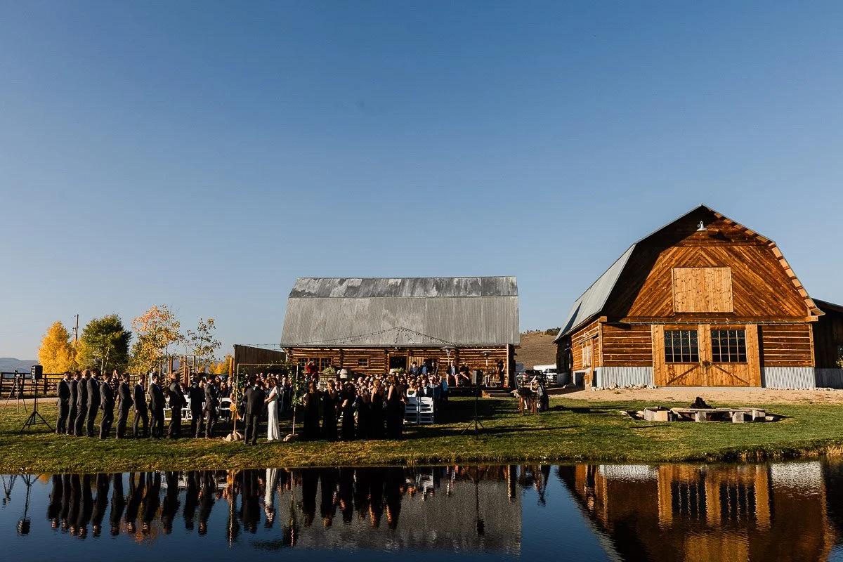 Outdoor wedding at a rustic ranch, with guests seated near wooden barns under a clear blue sky. Reflections shimmer on a calm pond, creating a serene ambiance during a Strawberry Creek Ranch wedding in Granby, Colorado