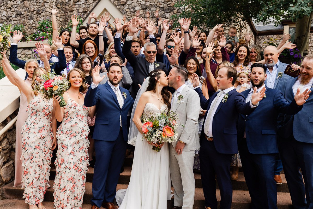 A joyful wedding scene with a bride and groom kissing in the center, surrounded by cheering guests. The crowd is waving and smiling, conveying celebration.