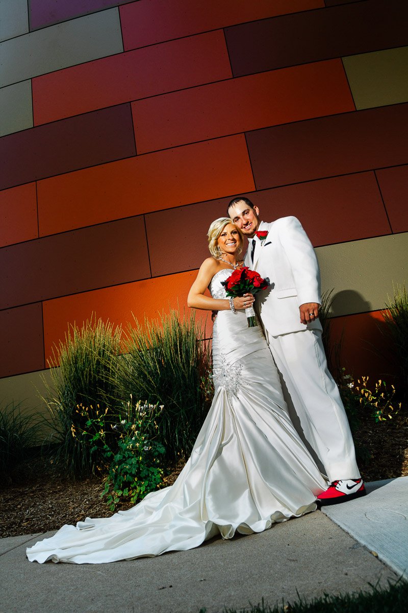 A smiling bride and groom pose for a portrait during a Lincoln Center wedding in Fort Collins. She's in a white dress holding red roses, and he wears a white suit with red accents. They're standing on a path with tall grass and a colorful, geometric 