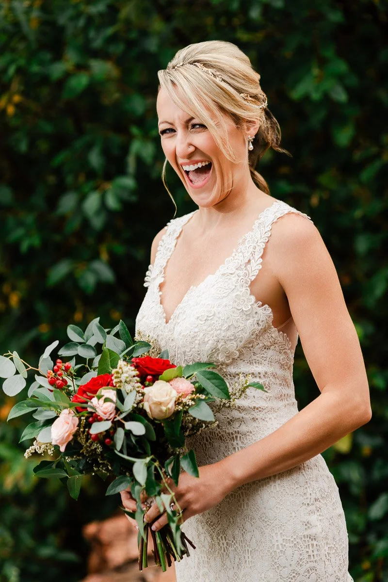 A joyful bride in a lace gown holds a vibrant bouquet of red and pink flowers with greenery. She is laughing, with a lush, green background.