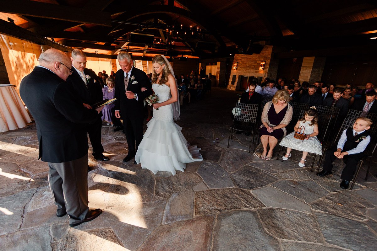 A bride and groom stand with an officiant and groomsman in a warmly lit, rustic wedding venue. Guests, including children, watch the ceremony.