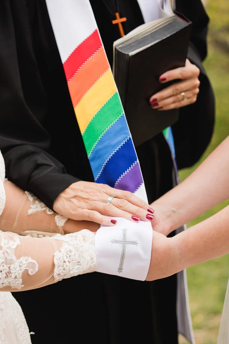 A minister with a rainbow sash and black robe holds hands of two people in wedding attire, one wearing a cross-embroidered sleeve. The scene conveys a sense of unity and support.