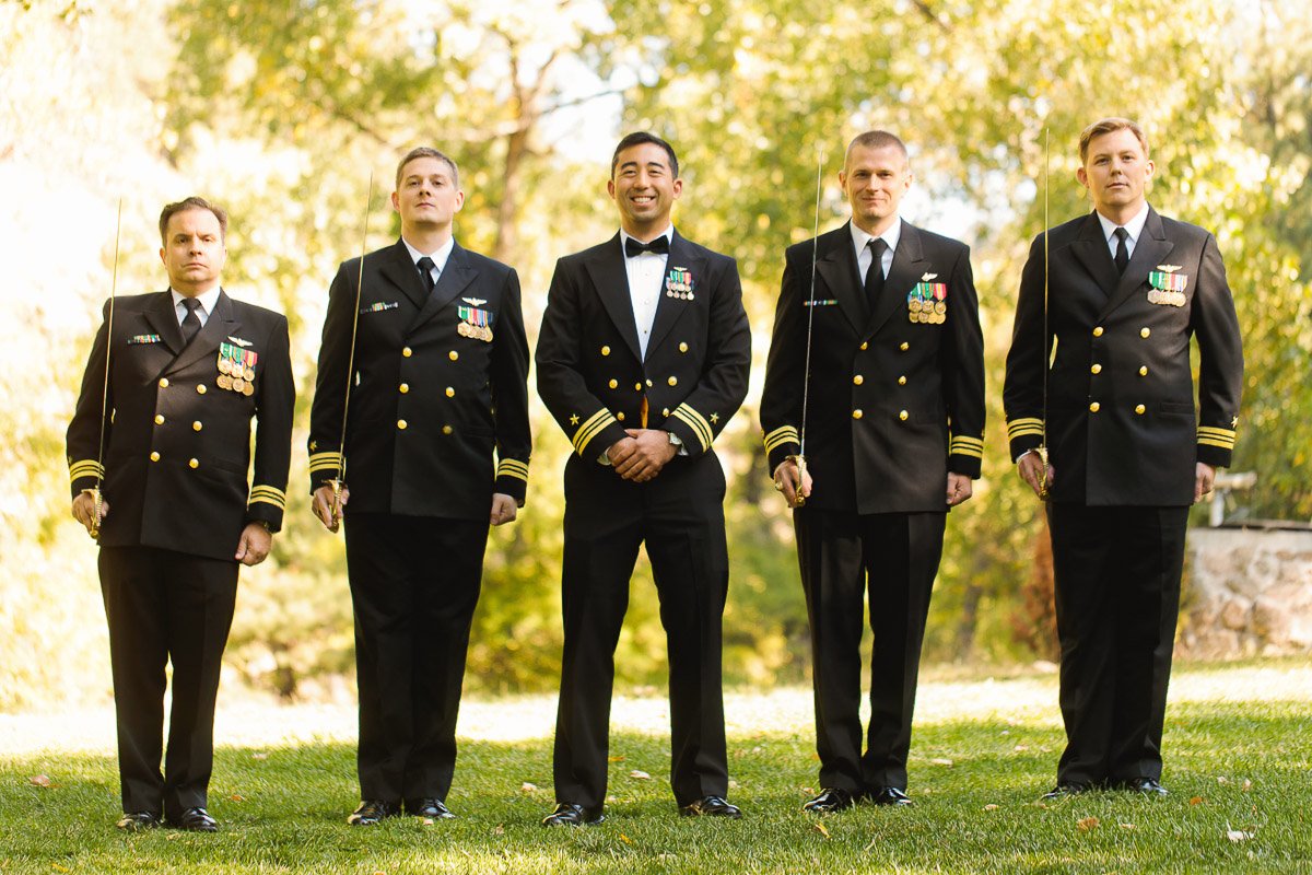 Five men in black naval uniforms with gold buttons and insignias stand on grass, smiling. Sunlight filters through green trees, creating a warm, formal atmosphere.