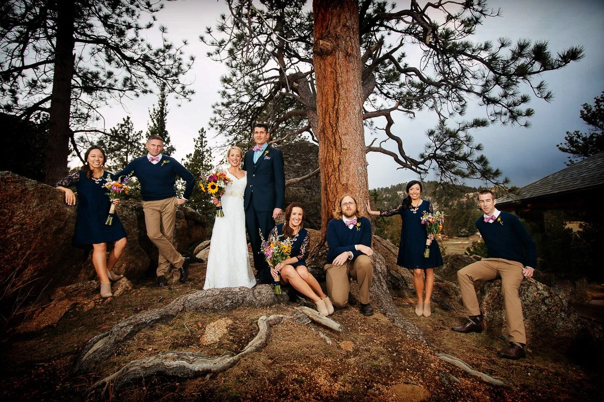 A wedding party poses outdoors under a large pine tree. The bride, in a white dress, stands with the groom, surrounded by smiling bridesmaids and groomsmen in blue and tan attire, each holding bouquets. The mood is joyful and celebratory.