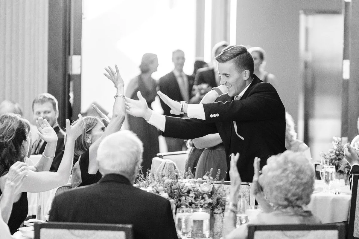 A man in a suit joyfully gives high-fives to seated guests at a formal event. The scene is lively and celebratory, with people smiling and engaged.