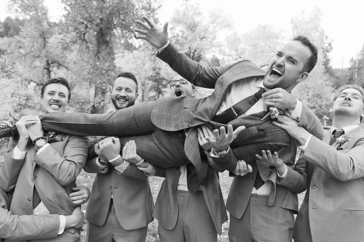 A group of smiling groomsmen in suits joyfully lift the groom in a playful pose outdoors. The black and white image conveys celebration and camaraderie.