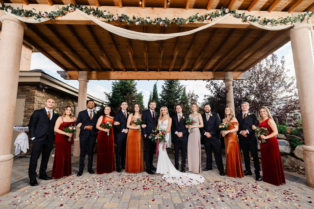 A wedding party stands under a floral pergola. The bride in a white gown is surrounded by bridesmaids in red and orange dresses and groomsmen in suits.