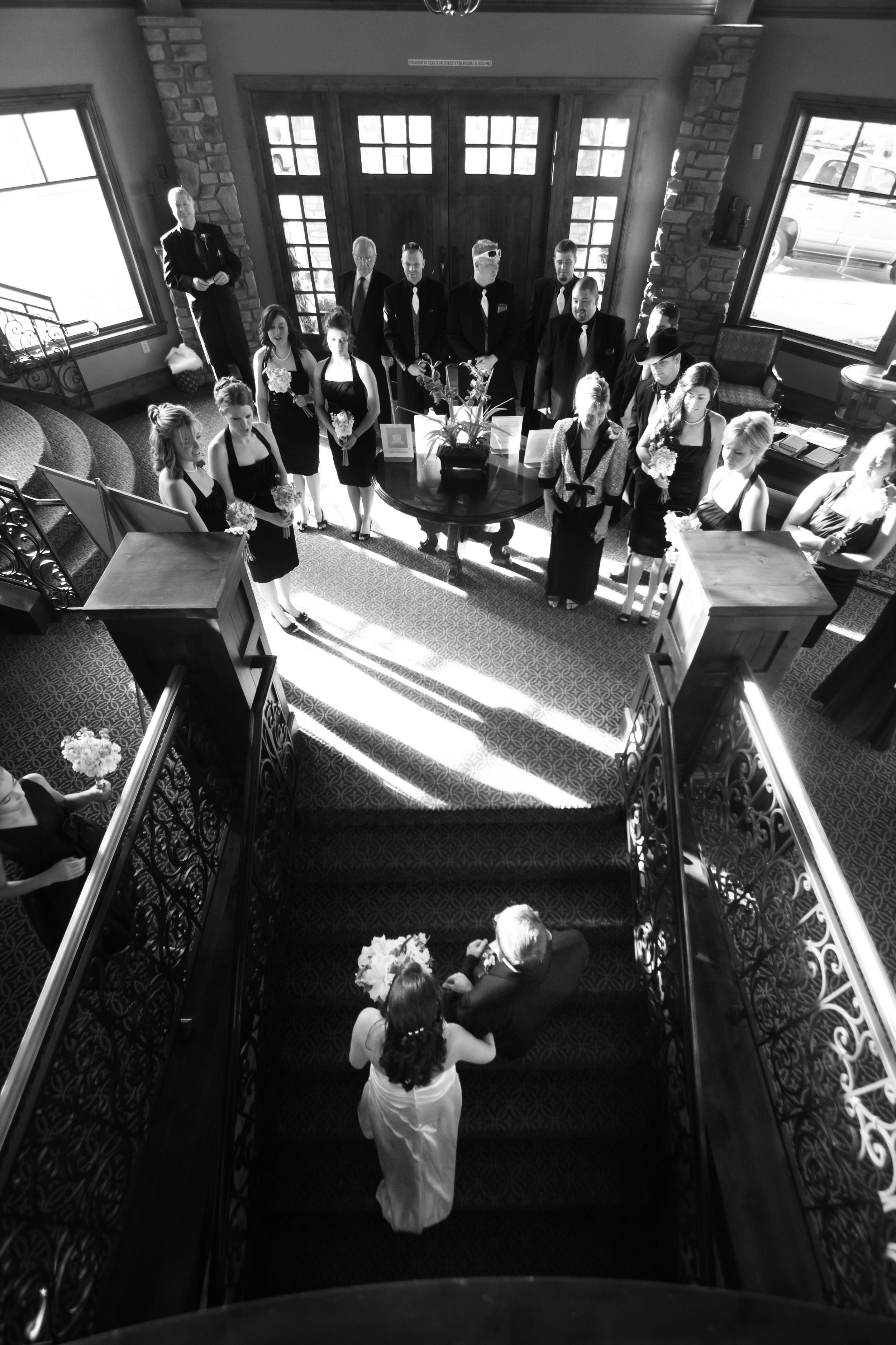Black and white image of a bride and father ascending a grand staircase towards an elegantly dressed wedding party above, illuminated by sunlight.