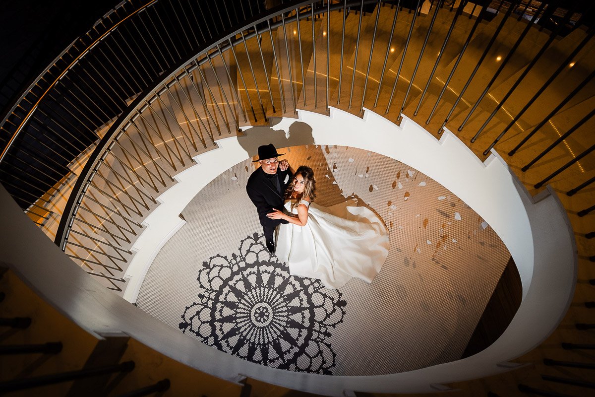 A bride and groom stand on a patterned floor in the center of a spiral staircase. The groom wears a black suit and hat, while the bride wears a flowing white gown.