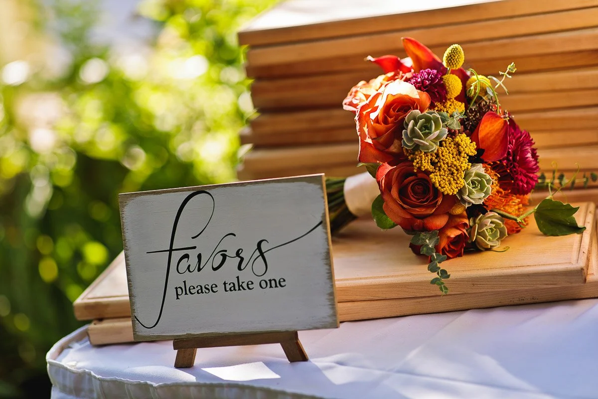 A wooden sign reading "favors please take one" stands on a table with stacked wooden boxes. Nearby is a vibrant bouquet with orange and red flowers.