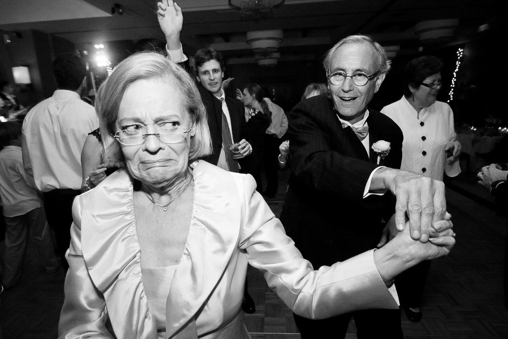 A black-and-white photo of an elderly couple dancing. The woman looks amused yet skeptical, while the man grins. Guests cheer in the background at a lively party captured by Colorado Wedding Photographer tomKphoto