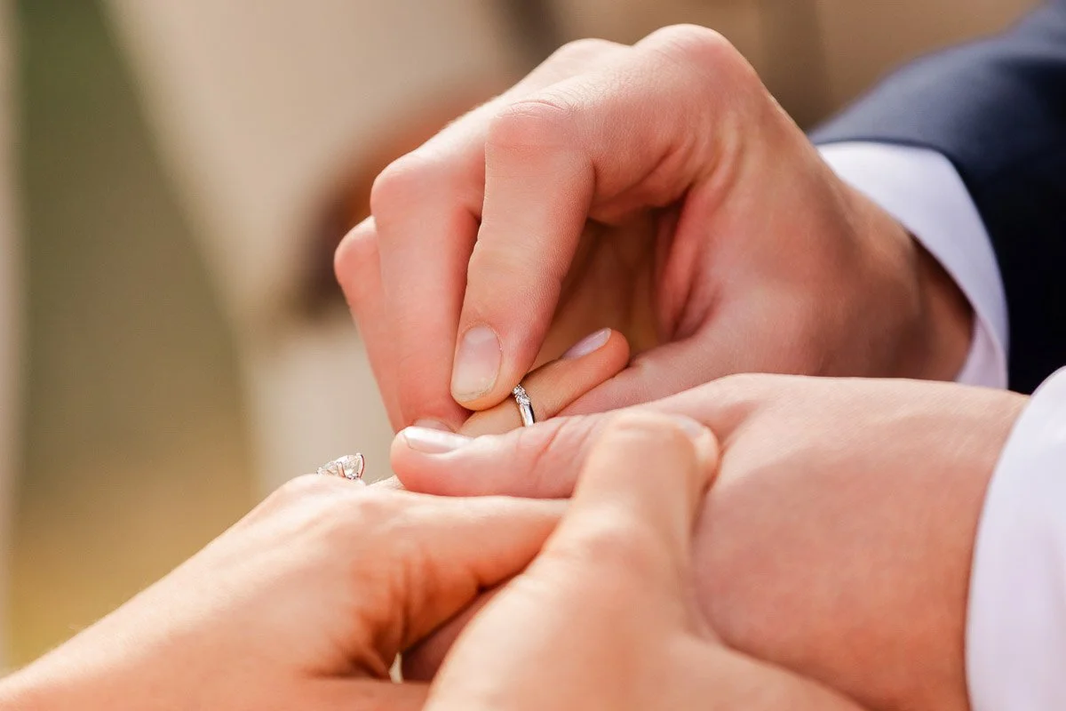 Close-up of a person placing a wedding ring on another's finger, conveying love and commitment. Hands are gently clasped, set against a soft background.