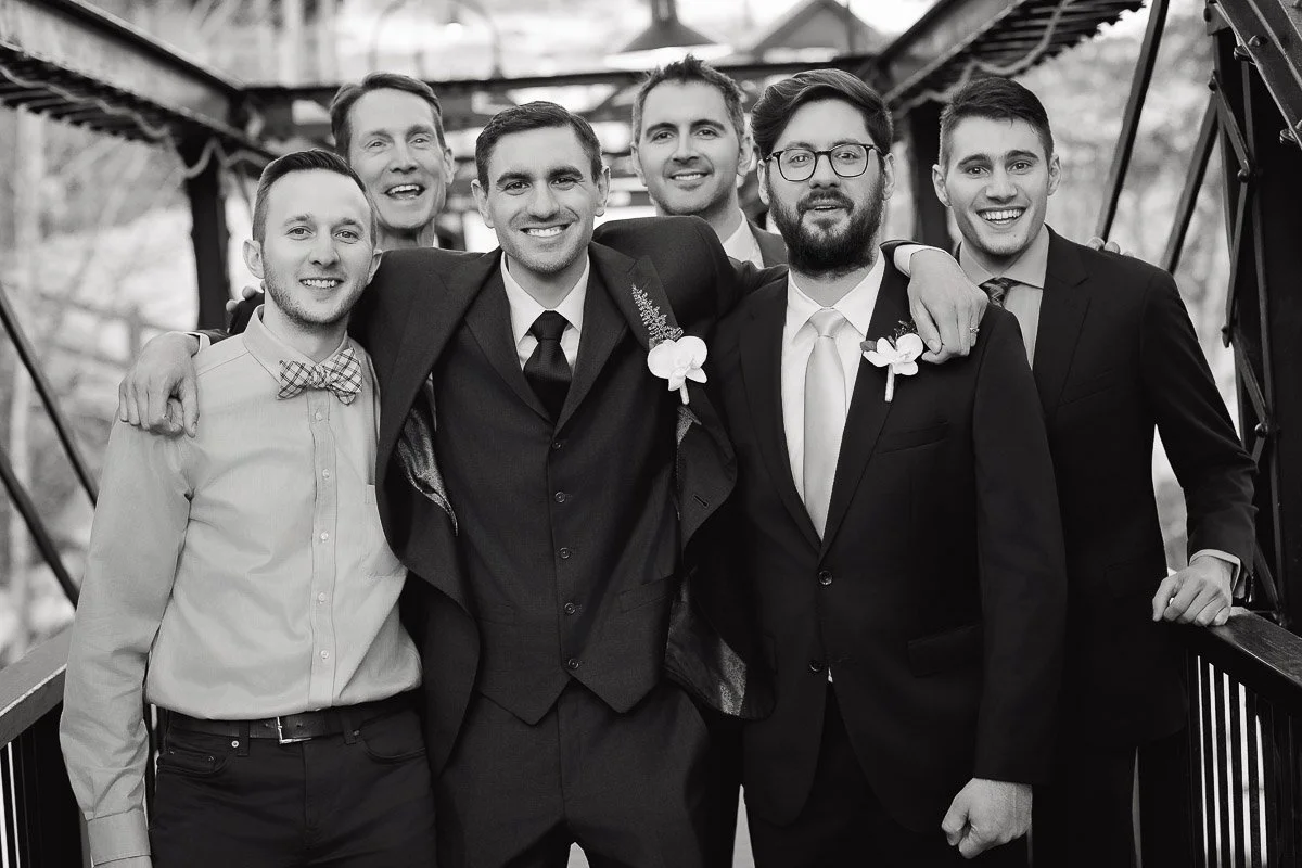 A group of six cheerful men pose on a metal bridge. Five wear suits with boutonnières, and one wears a shirt with a bow tie. The tone is joyful and celebratory.