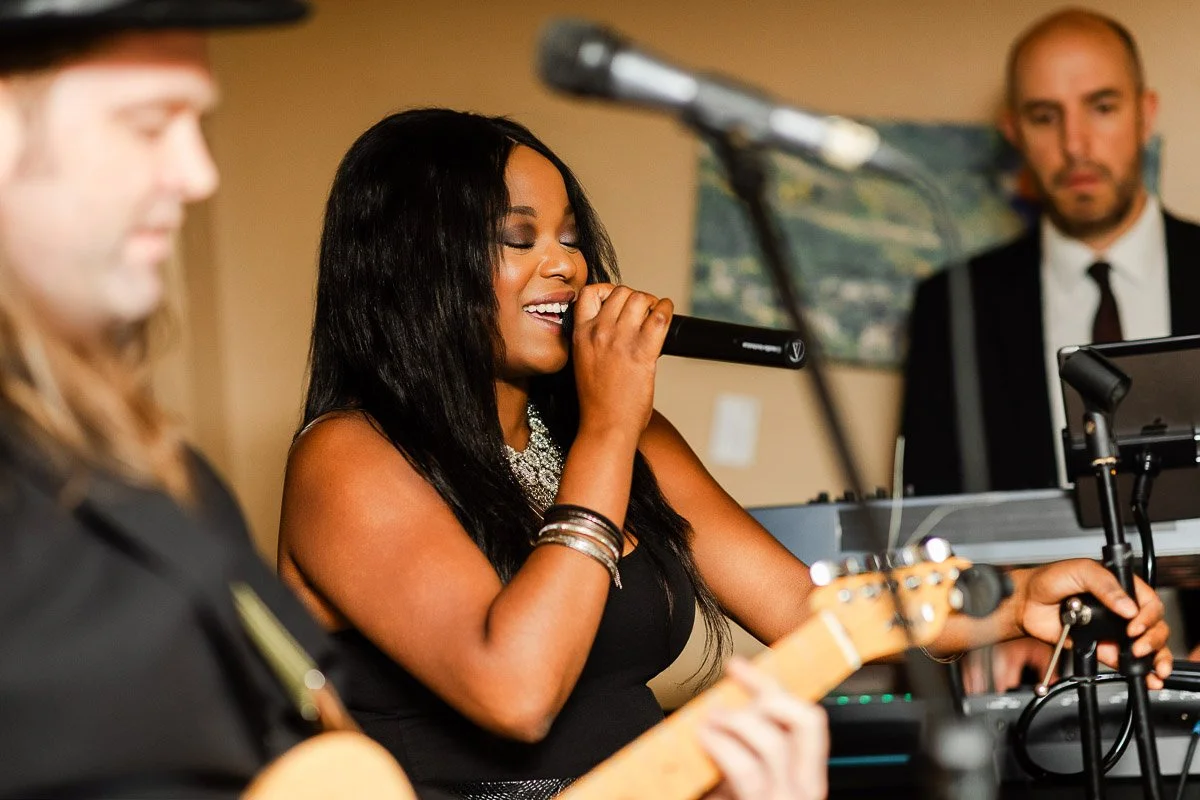 A woman passionately sings into a microphone on stage, surrounded by musicians playing instruments. The atmosphere is lively and vibrant.