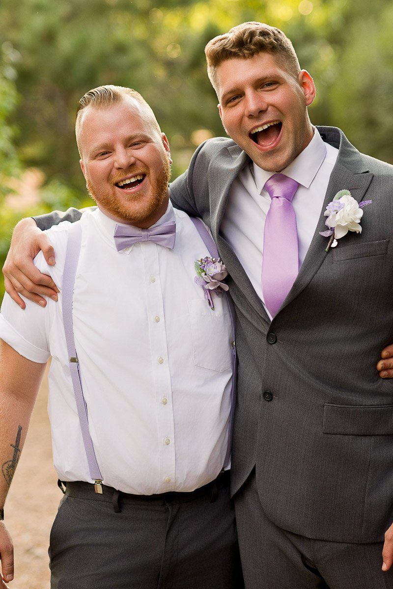 Two joyful men in formal attire, one in a gray suit with a purple tie, and the other in a white shirt, purple bow tie, and suspenders, smile and embrace outdoors.