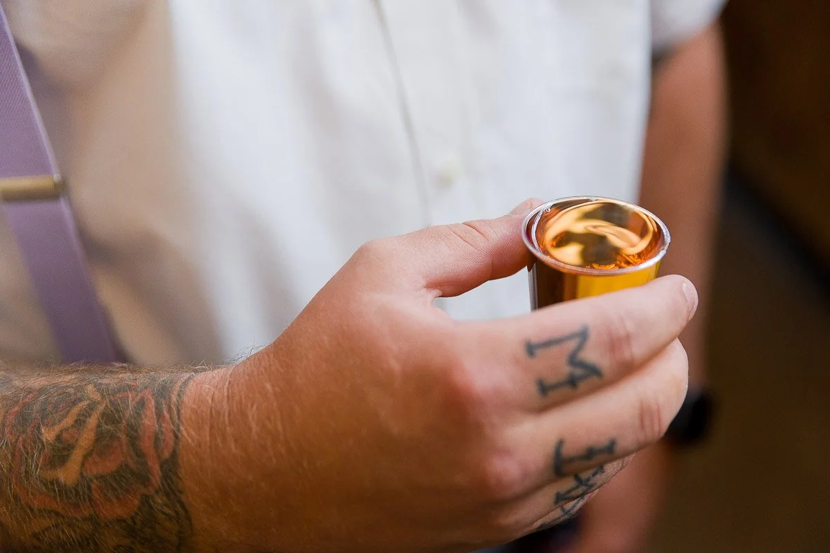 A person with tattooed fingers holds a small gold cup of espresso. They wear a white shirt and lavender suspenders. The mood is calm and casual.
