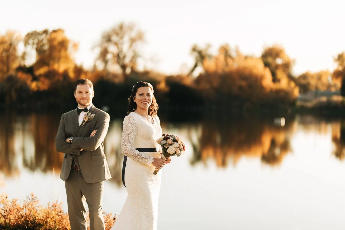 A bride in a white lace dress holds a bouquet by a calm lake during a Wellshire Event Center wedding, with a groom in a gray suit beside her. Autumn trees reflect on the water. Serene and joyful.