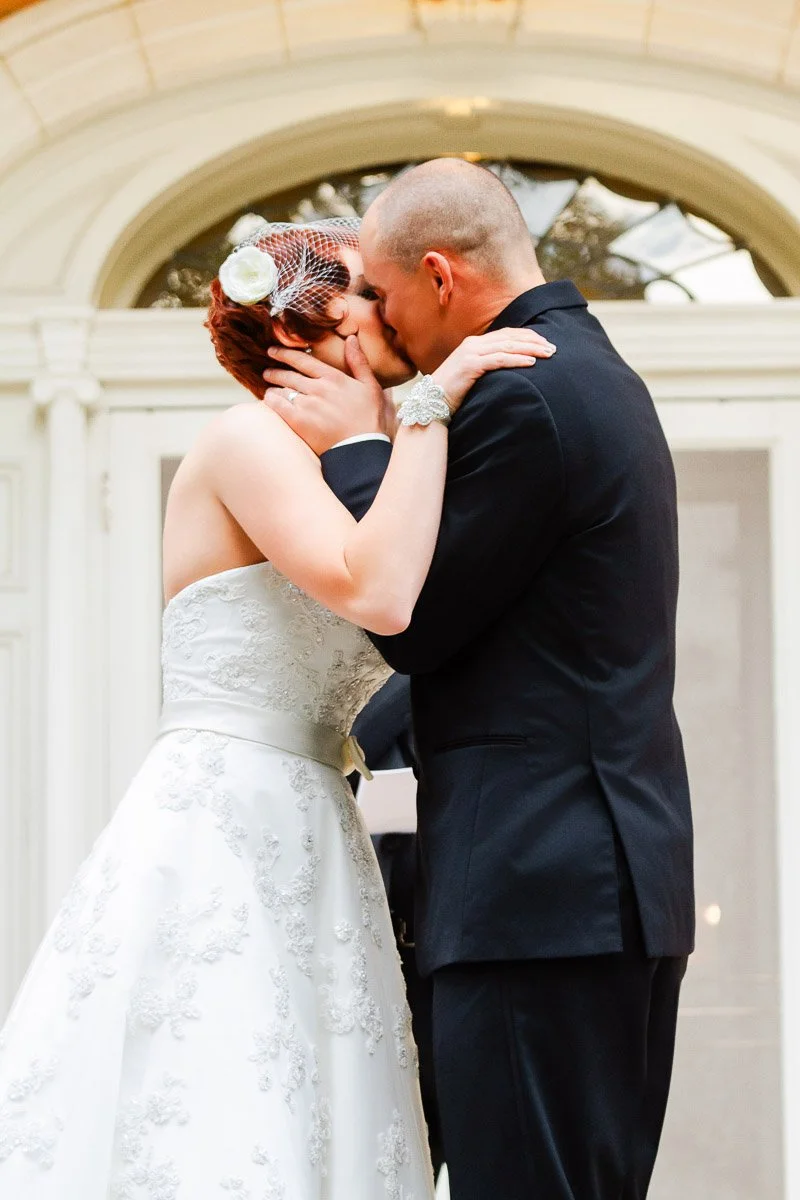 A couple shares a kiss at their wedding. The bride wears a white gown with floral embroidery and a birdcage veil, and the groom is in a black suit. The backdrop is an elegant, arched doorway, conveying romance and joy.