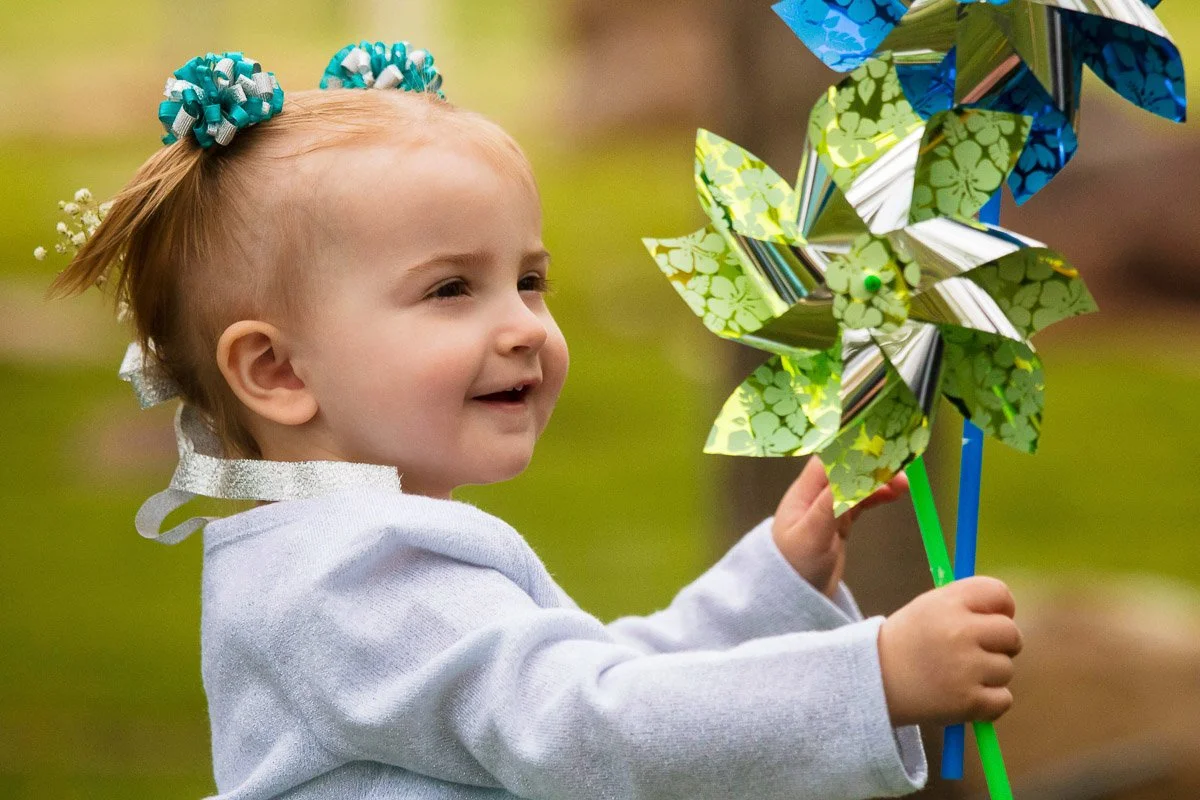 A young child with floral hair ties holds colorful pinwheels, smiling joyfully outdoors. The scene conveys innocence and playfulness.