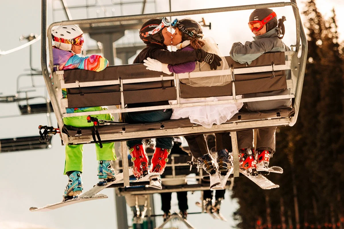 Couple in wedding attire and ski gear kiss on a ski lift, flanked by two skiers in colorful outfits. The scene is joyful and lighthearted, captured by Colorado Wedding Photographer, tomKphoto