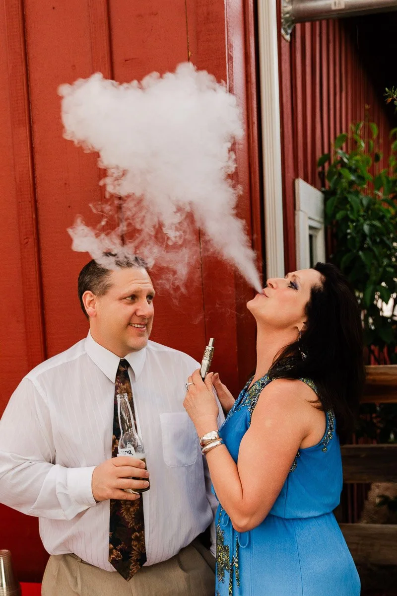 A woman in a blue dress exhales vapor forming a heart shape, smiling, while a man in a white shirt with a drink stands nearby. They are outdoors by a red wall.