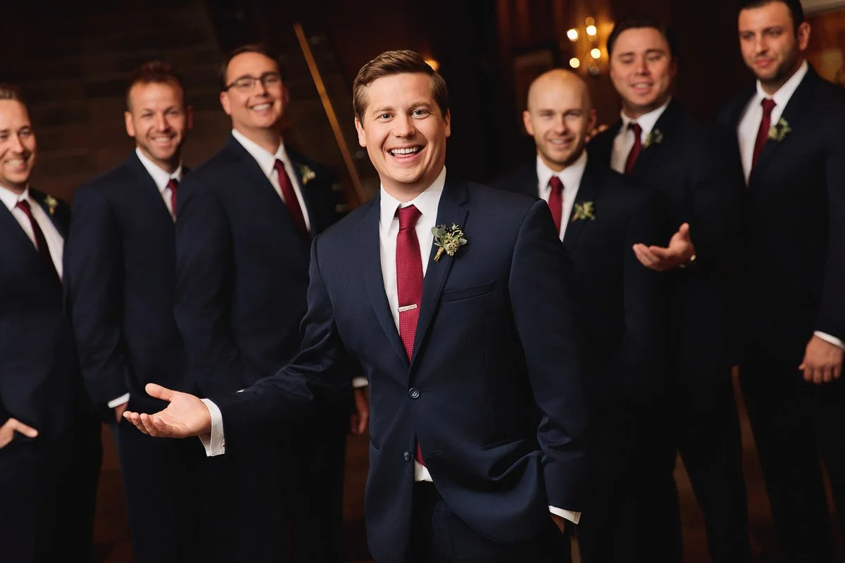 A smiling groom in a dark suit with a red tie stands with his groomsmen in a warm-lit venue. All wear matching attire, conveying joy and camaraderie.
