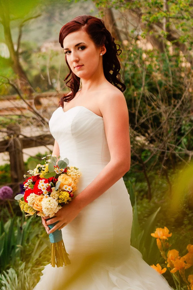 Bride in a strapless white gown, holding a vibrant bouquet of red, yellow, and white flowers. Standing in a lush garden, she looks serene and composed.