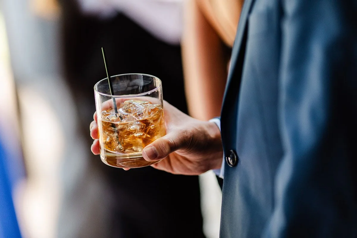 A person in a suit holds a glass of whiskey with ice and a black straw, suggesting a sophisticated social setting. The background is softly blurred.